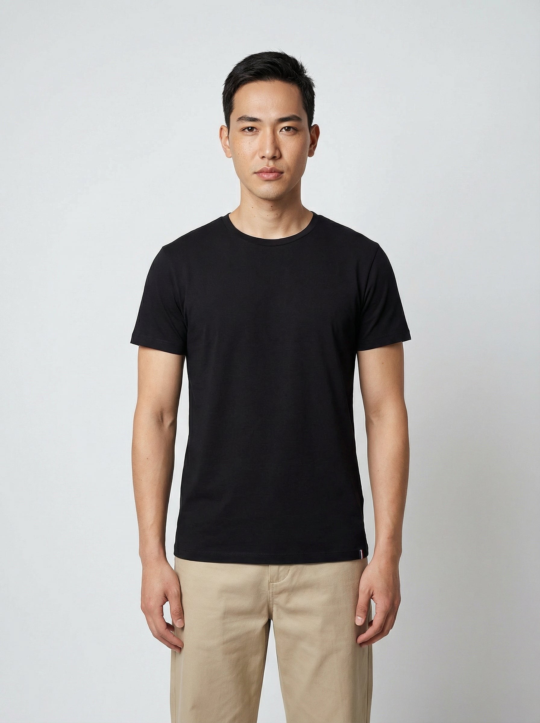 Homme en t-shirt noir et pantalon beige, debout sur fond gris clair